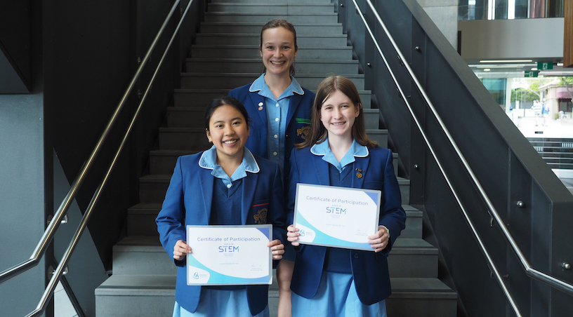 Khanh, Lucy, and Jemima standing on staircase holding their certificates